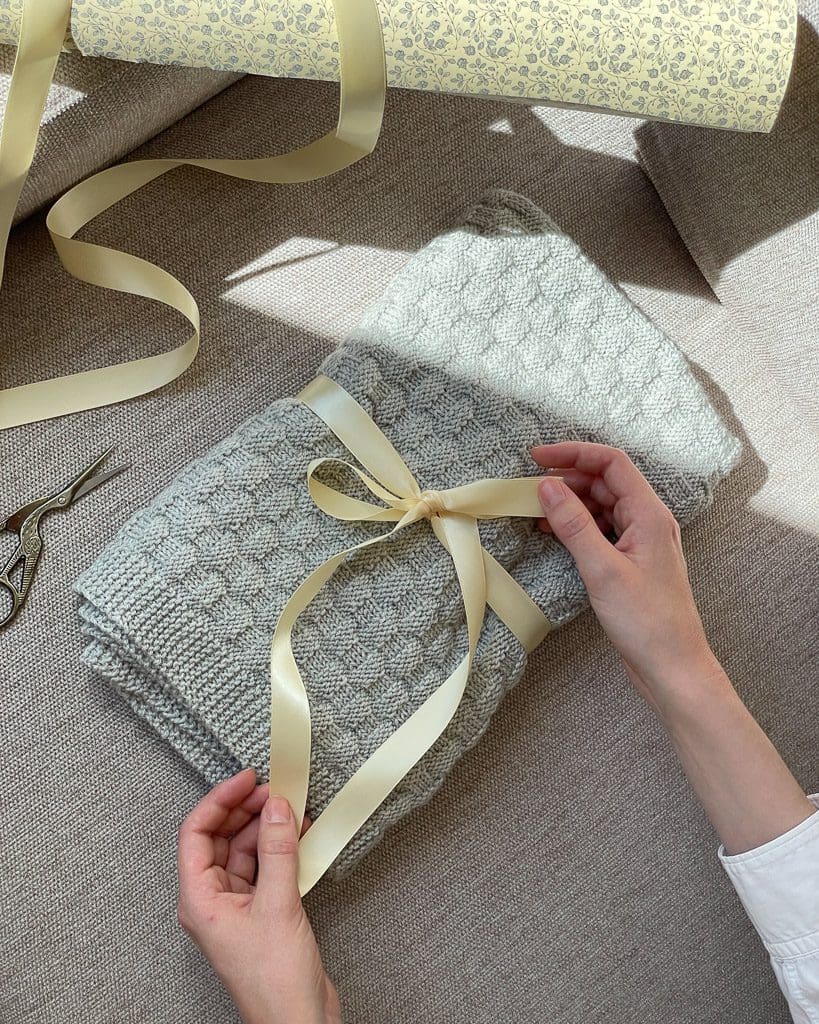 En person er ved at pakke et lysegrå meleret Chess Baby Blanket i det fineste ternede mønster.