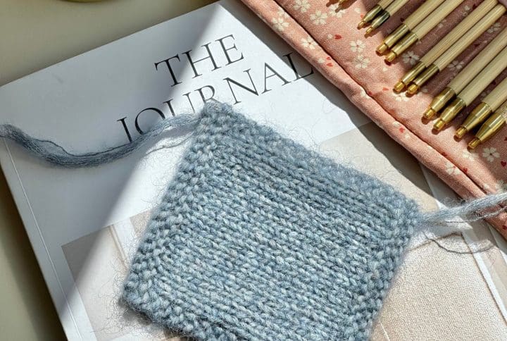 En strikkeprøve til at måle strikkefastheden med. Den ligger på en bog i sollyset. I baggrunden er et pindesæt fra Seeknit.