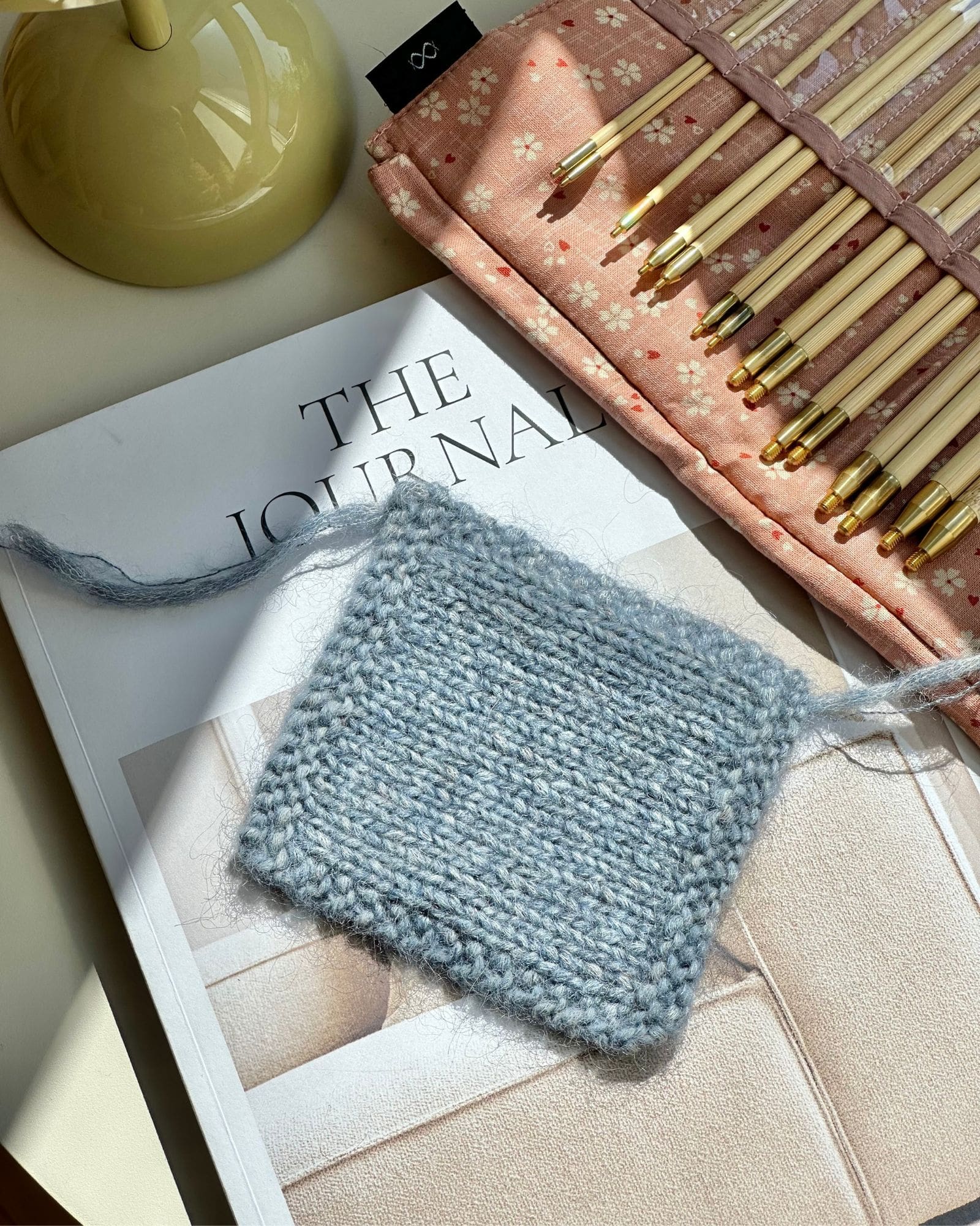 En strikkeprøve til at måle strikkefastheden med. Den ligger på en bog i sollyset. I baggrunden er et pindesæt fra Seeknit.